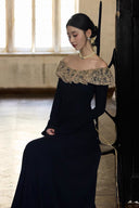 Seated view of black off-shoulder maxi dress with antique lace ruffle neckline and fitted long sleeves