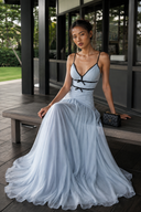 Seated view of the Vogsion Célestine light blue wedding guest dress with black bow details, fitted bodice, and airy flowing maxi skirt on a modern resort veranda.
