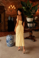 Rear view of the Vogsion Cyra Asymmetrical Draped Maxi Dress highlighting the lace-up corset back, sculpted waist and softly draped butter yellow maxi skirt.