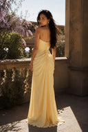 Back view of the Vogsion Cyra Asymmetrical Draped Maxi Dress featuring a lace-up corset back, gathered draping and butter yellow maxi skirt in a Mediterranean-inspired outdoor setting.