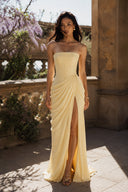 Vogsion Cyra Asymmetrical Draped Maxi Dress in butter yellow featuring a strapless corset bodice, asymmetrical draping and thigh-high slit, photographed outdoors in warm natural sunlight.