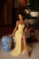 Model wearing the Vogsion Cyra Asymmetrical Draped Maxi Dress in butter yellow while seated indoors, showcasing the strapless corset, asymmetrical draping and elegant thigh-high slit.