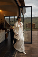 Full-length side view of an ivory plunge neck maxi dress styled with an ivory coat and handbag