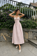 Woman in a Vogsion Roselle Blush Floral Corset Lace-Trim Midi Dress standing outdoors with greenery and a stone wall in the background