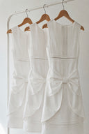 Three Vogsion Shushu White Linen Statement Bow Petite Midi Dresses hanging on wooden hangers. Premium white linen petite midi dress featuring pintuck detailing, lace trim, oversized waist bow, sculptural layered skirt, and a timeless minimalist design for summer, bridal events, and vacation wear