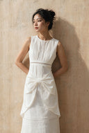 Vietnamese high-cheekbone model wearing the Vogsion Shushu White Linen Statement Bow Petite Midi Dress in a warm textured studio setting. White linen petite midi dress with delicate pintuck lace panel, oversized statement bow at the waist, sculptural draped overlay, and breathable natural fabric, photographed in soft spring daylight.
