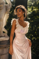 Full-length view of the Vogsion Sunset Aurelia Lace-Trimmed Liquid Satin Gown featuring shimmering blush liquid satin, lace-trimmed neckline, flowing draped skirt, and romantic garden backdrop. Luxury wedding guest maxi dress, formal satin gown, cottagecore evening dress, fairycore bridesmaid outfit, elegant summer occasion wear.