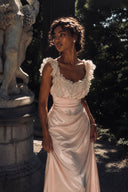 Model wearing the Vogsion Sunset Aurelia Lace-Trimmed Liquid Satin Gown in blush pink with delicate lace ruffle straps and a draped liquid satin silhouette, posing beside a classical garden statue in warm golden sunlight. Elegant wedding guest dress, romantic evening gown, luxury satin maxi dress, fairycore formalwear.