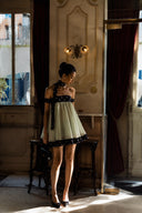 Full-body editorial image of Vogsion Velvandis Off Shoulder Dot Mini Dress (Sage Green Polka Dot) featuring an elegant off-shoulder neckline, soft gathered skirt, contrast polka dot hem and matching scarf. A feminine mini dress for wedding brunches, European vacations, garden parties and luxury summer styling.
