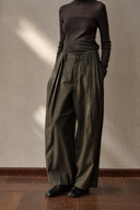 Close-up of olive wool-blend trousers showing the high waist, front pleats and curved wide legs.